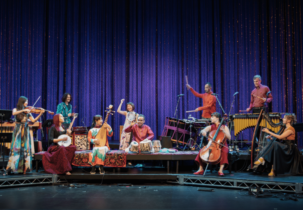 Image of a collective of artists dressed in colorful clothing with various instruments. They are gathered around on stage for a performance. 
